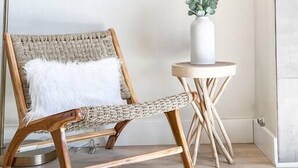 Ein Zimmer mit einem Holzstuhl, einem kleinen weißen Tisch mit einer Pflanzvase und einer Stehlampe.