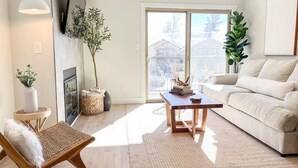 Ein modernes Wohnzimmer mit einer Couch, einem Holztisch und einem Sessel. Ein Kamin, ein an der Wand befestigtes Fernsehgerät und ein großes Fenster mit Blick auf Häuser sind vorhanden.