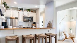 Eine moderne Küche mit weißen Schränken, einer Edelstahl-Mikrowelle und einer Holz Arbeitsplatte. Drei bequeme Barhocker aus Holz sind ebenfalls vorhanden.