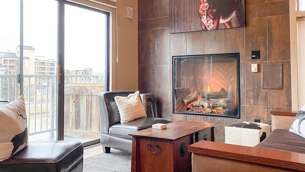 A modern living room with a fireplace, a leather sofa, and a large window with a view of buildings. - Slopeside Chalet (Steamboat Springs)