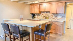A kitchen with wooden cabinets, a white countertop, and a tiled backsplash. There are two black chairs and a white island with a glass vase. - Slopeside Chalet (Steamboat Springs)