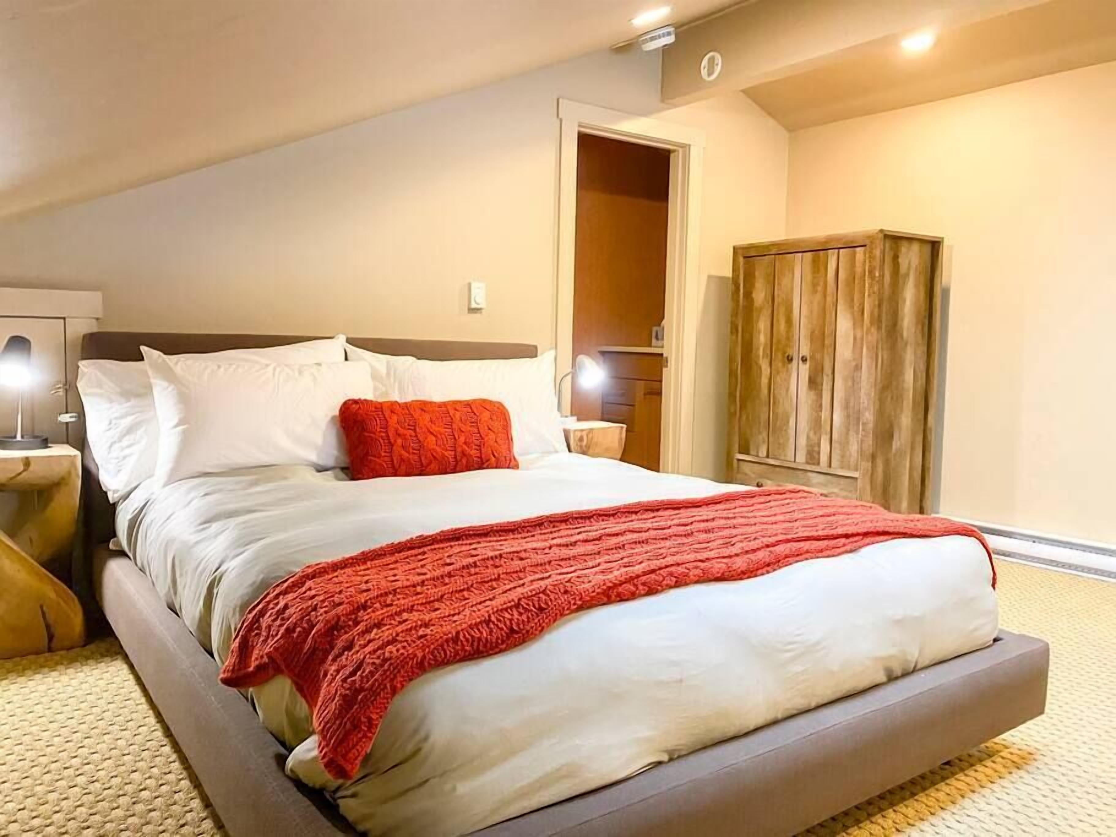 Een opgemaakt bed met wit en rode beddengoed, een houten kledingkast en een bedlampje.