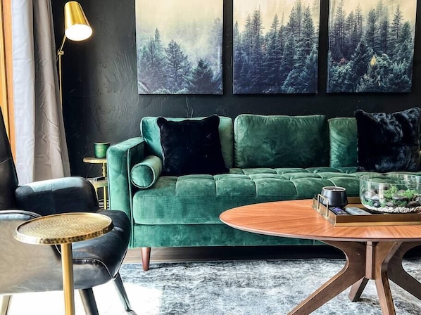 A modern living room with a green sofa, a round wooden coffee table, and a black armchair. There are three paintings of forest scenes on the wall. - Evergreen Escape Summ (Steamboat Springs)