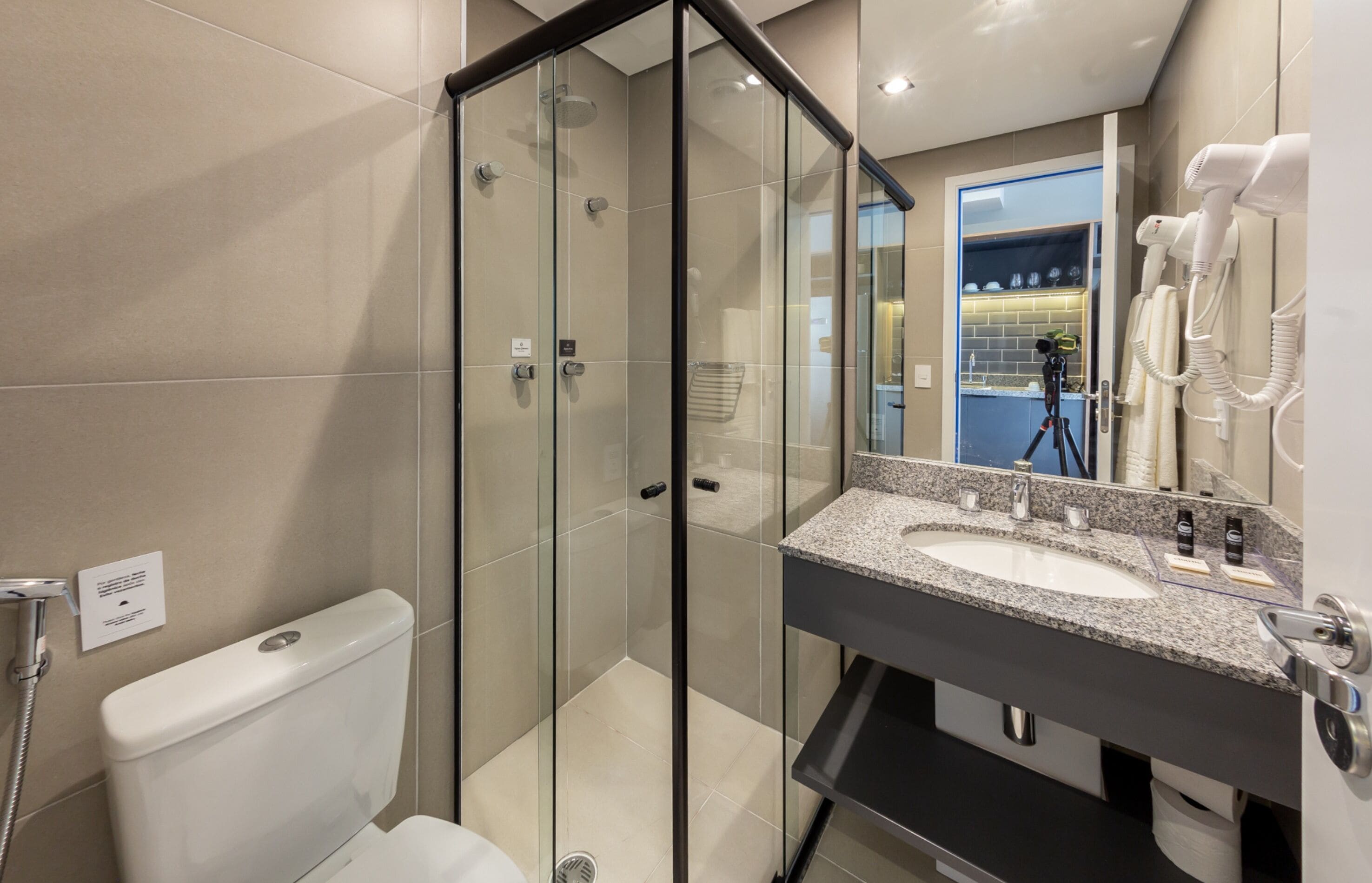 Une salle de bain moderne avec une douche à parois de verre, des toilettes blanches, un lavabo surmonté d’un plan de travail en granit et un sèche-cheveux fixé au mur.