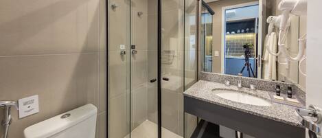 A modern bathroom with a glass-enclosed shower, a white toilet, a granite countertop sink, and a hairdryer mounted on the wall.