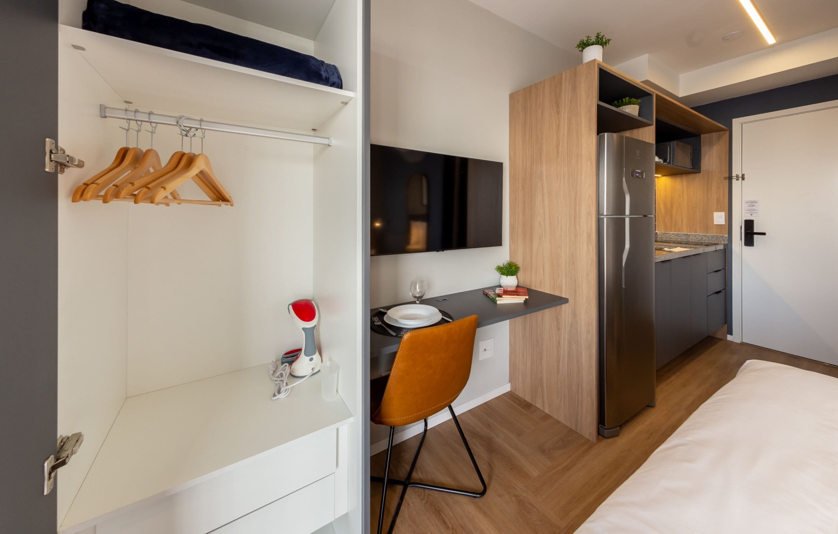 Ein kompaktes Hotelzimmer mit Küchenzeile, einem Bett, einem Schreibtisch mit Stuhl und einem Kleiderschrank mit Kleidung.