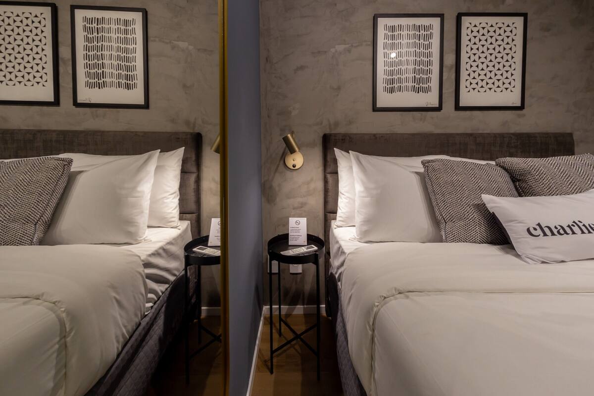 A hotel room with two beds, a bedside table, a wall-mounted lamp, and framed artwork on the wall.