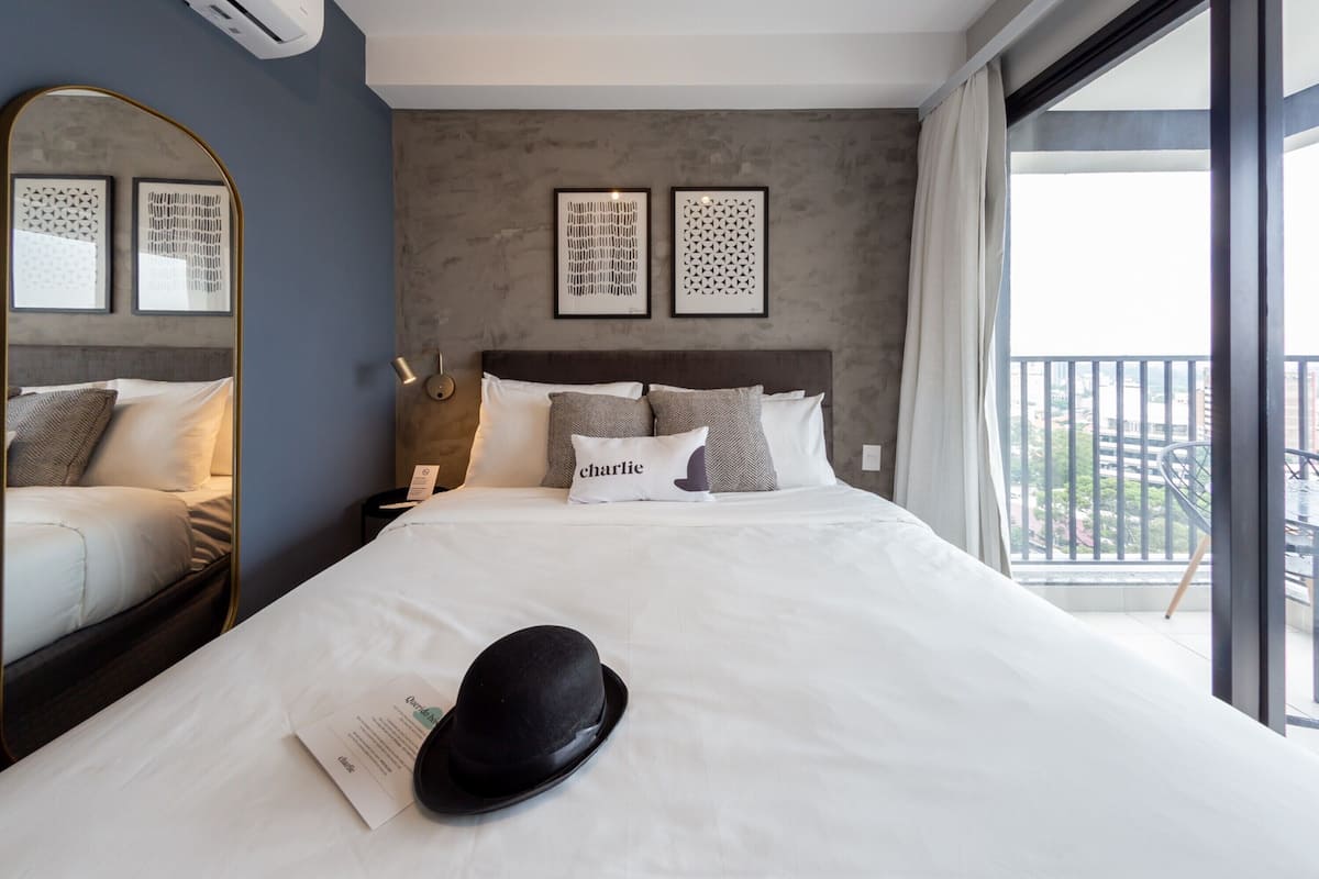 A neatly made bed with a black hat and a pillow with the name "Charlie" on a hotel room with a large mirror, framed artwork, and a balcony with a view.