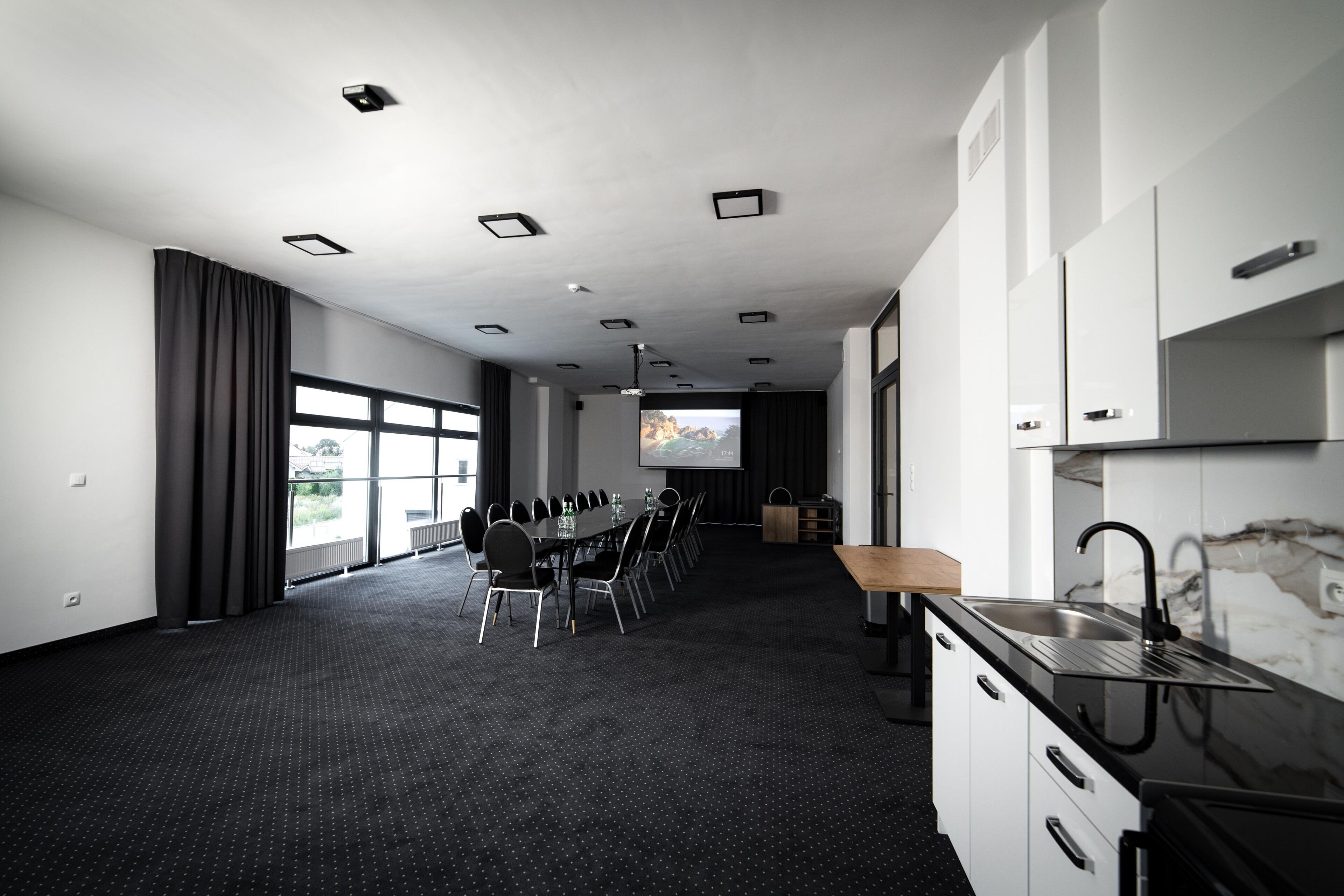 Un salón de conferencias con una larga mesa, sillas, una pantalla de proyector y una zona de cocina con un lavabo y armarios.