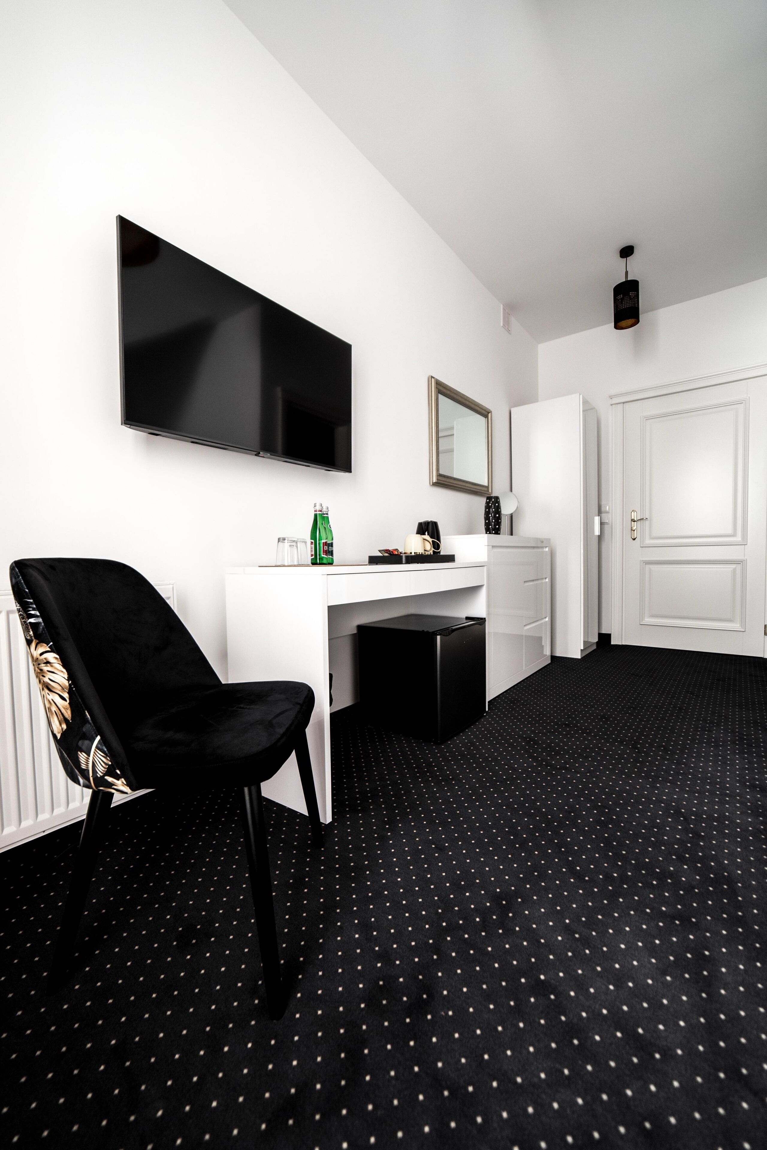 Ein Hotelzimmer mit einem weißen Schreibtisch, einem schwarzen Stuhl, einem flachbildfernseher an der Wand, einem kleinen Spiegel und einer Tür.