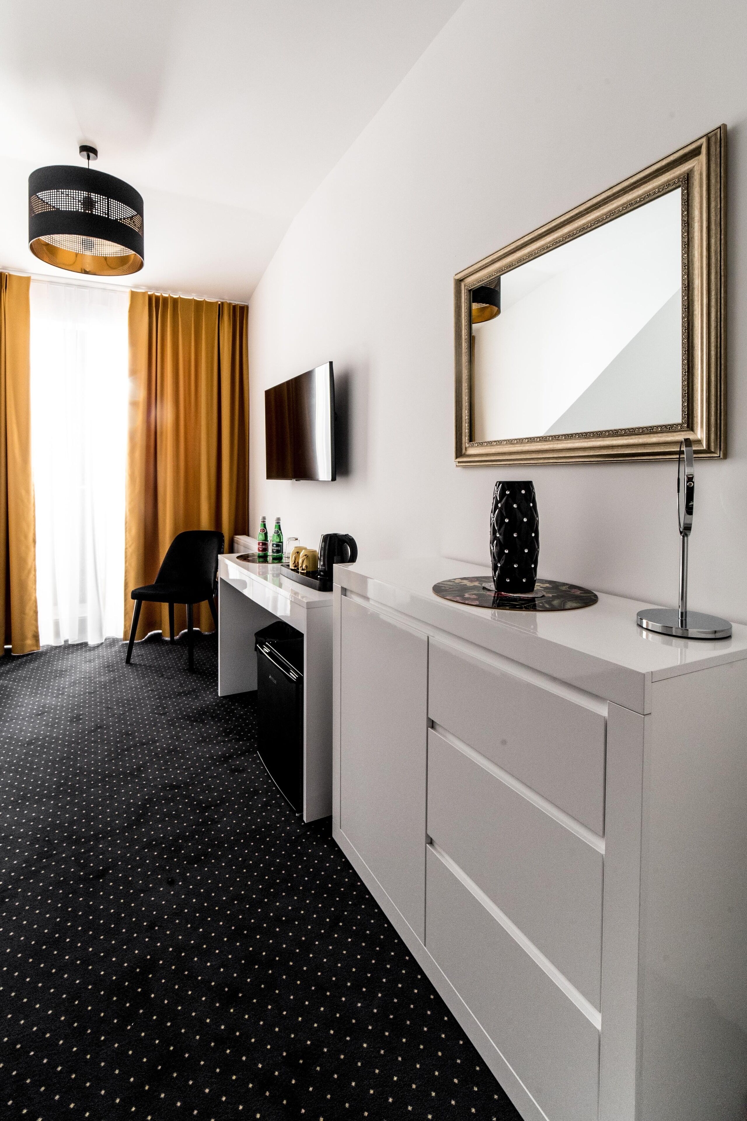 A hotel room with a white desk, a black chair, a flat-screen TV, a large mirror, a vase, and a coffee maker.