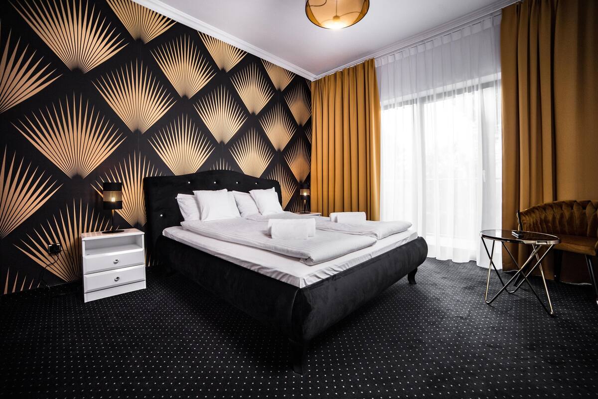 A modern hotel room with a large bed, a nightstand, a chair, and a patterned wall.