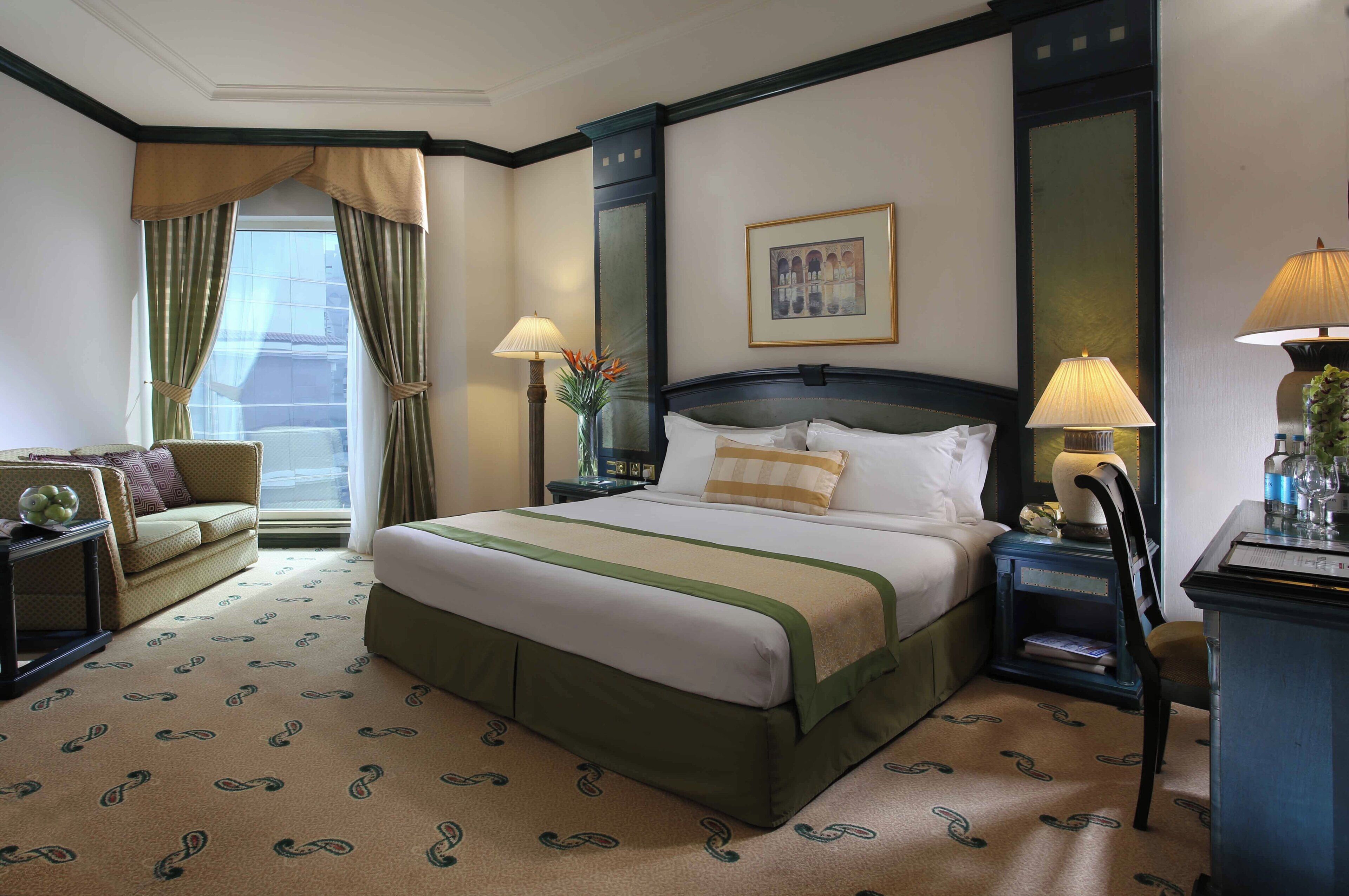 A hotel room with a large bed, a sofa, a desk with a lamp, and a window with curtains.