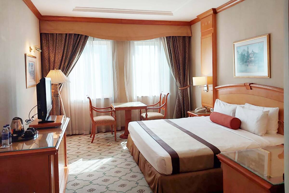 A hotel room with a bed, a desk, a chair, a table, and two windows with curtains.