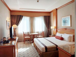 Une chambre d’hôtel comprenant un lit, un bureau, une chaise, une table et deux fenêtres avec des rideaux.