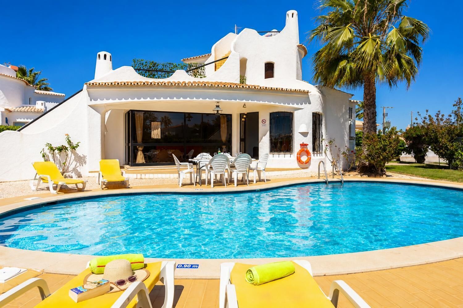 A poolside area with lounge chairs, a pool, and a building with a white facade and a palm tree.