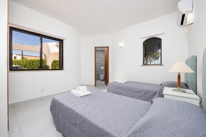 A hotel room with two beds, a window with a view of a building and greenery, a bathroom with a shower and toilet, and a bedside table with a lamp.