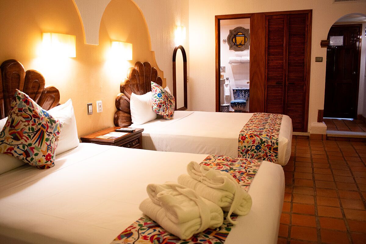 Habitación de hotel con dos camas, cabecera de madera, un espejo y un baño visible a través de una puerta abierta.