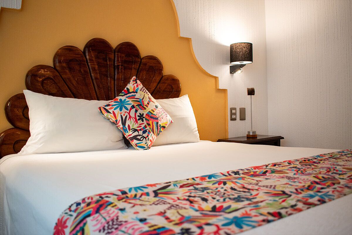 Een hotelkamer met een bed, een decoratief hoofdbord, een nachtkastje met een lamp en een wandlamp.