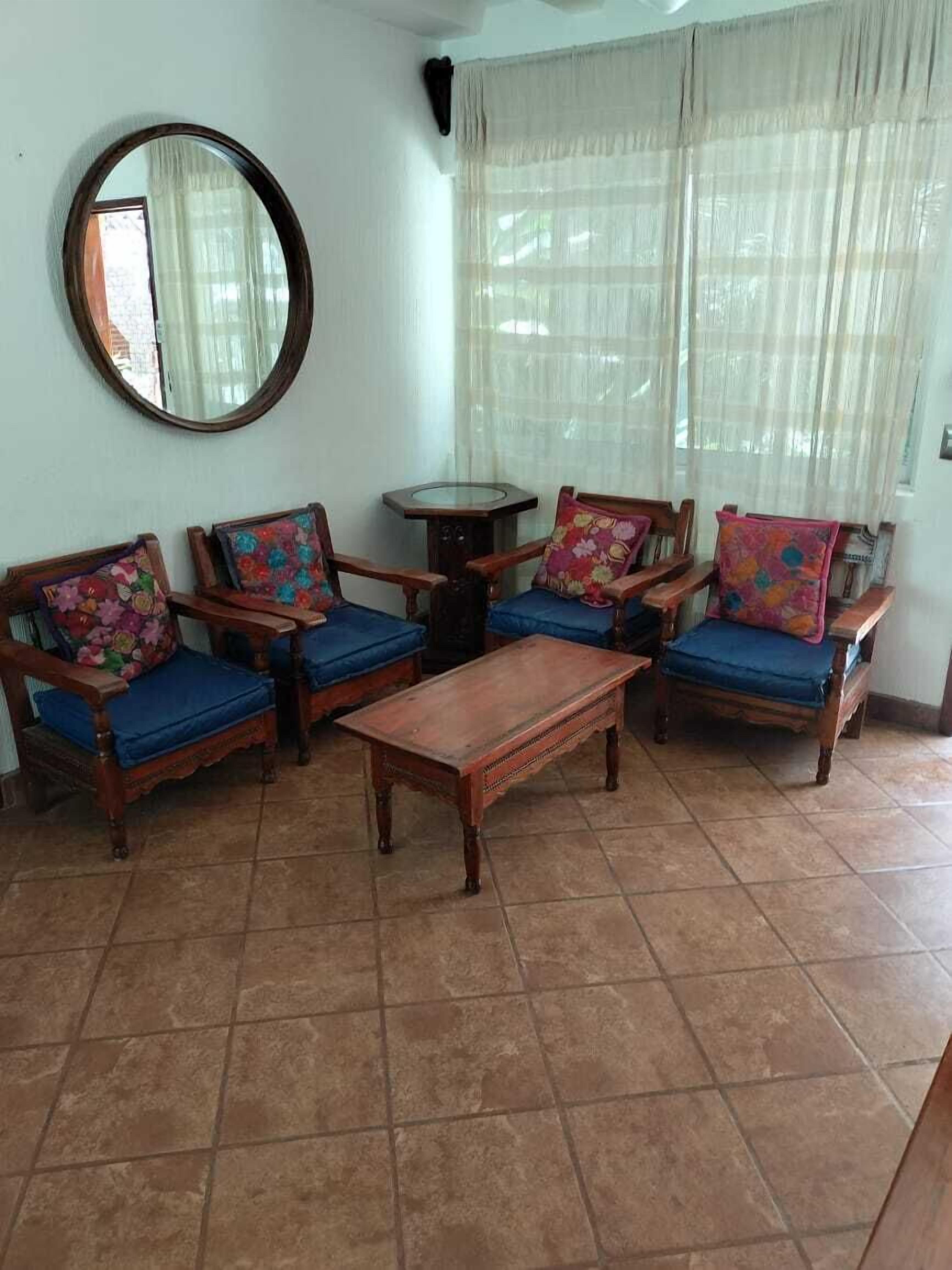 Una sala de estar con cuatro sillones de madera con cojines azules y almohadas estampadas, una mesa de centro de madera, un espejo redondo en la pared y una mesita redonda.