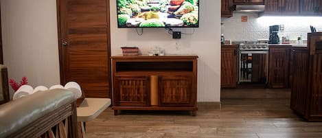 Un salon avec un meuble TV en bois, un téléviseur à écran plat fixé au mur, et un coin cuisine doté de meubles en bois et d’une cuisinière.