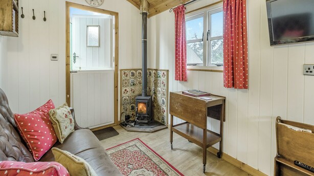 A cozy living room with a fireplace, wooden ceiling, and a television mounted on the wall. - Shepherds Hut - UK45529 (Leighton, near Shrewsbury)