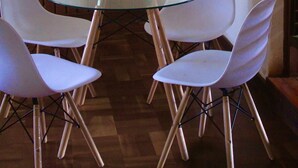 Una mesa redonda de vidrio con cuatro sillas blancas, un muro de ladrillo y un armario de madera.