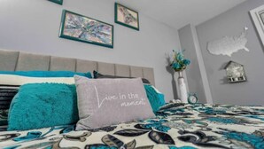 A bedroom with a bed, a pillow with the text "Live in the moment," and a wall with framed artwork.