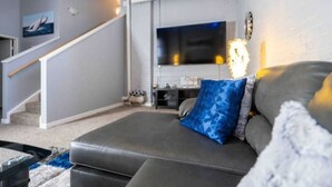 A modern living room with a grey sofa, a blue patterned pillow, a flat-screen TV mounted on the wall, and a staircase with a wooden railing.