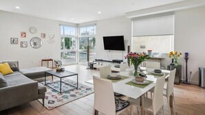 A modern living room with a sofa, coffee table, dining table, and chairs. There is a flat-screen TV mounted on the wall, a large window with a view of the outdoors, and a small side table with a vase of flowers. - Close to Hollywood and Getty with a Panorama Deck (Los Angeles)
