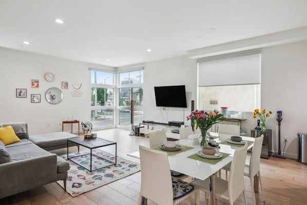 A modern living room with a sofa, coffee table, dining table, and chairs. There is a flat-screen TV mounted on the wall, a large window with a view of the outdoors, and a small side table with a vase of flowers.