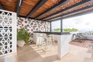 A rooftop terrace with a wooden ceiling, a geometric patterned wall, and a bar area with white chairs.