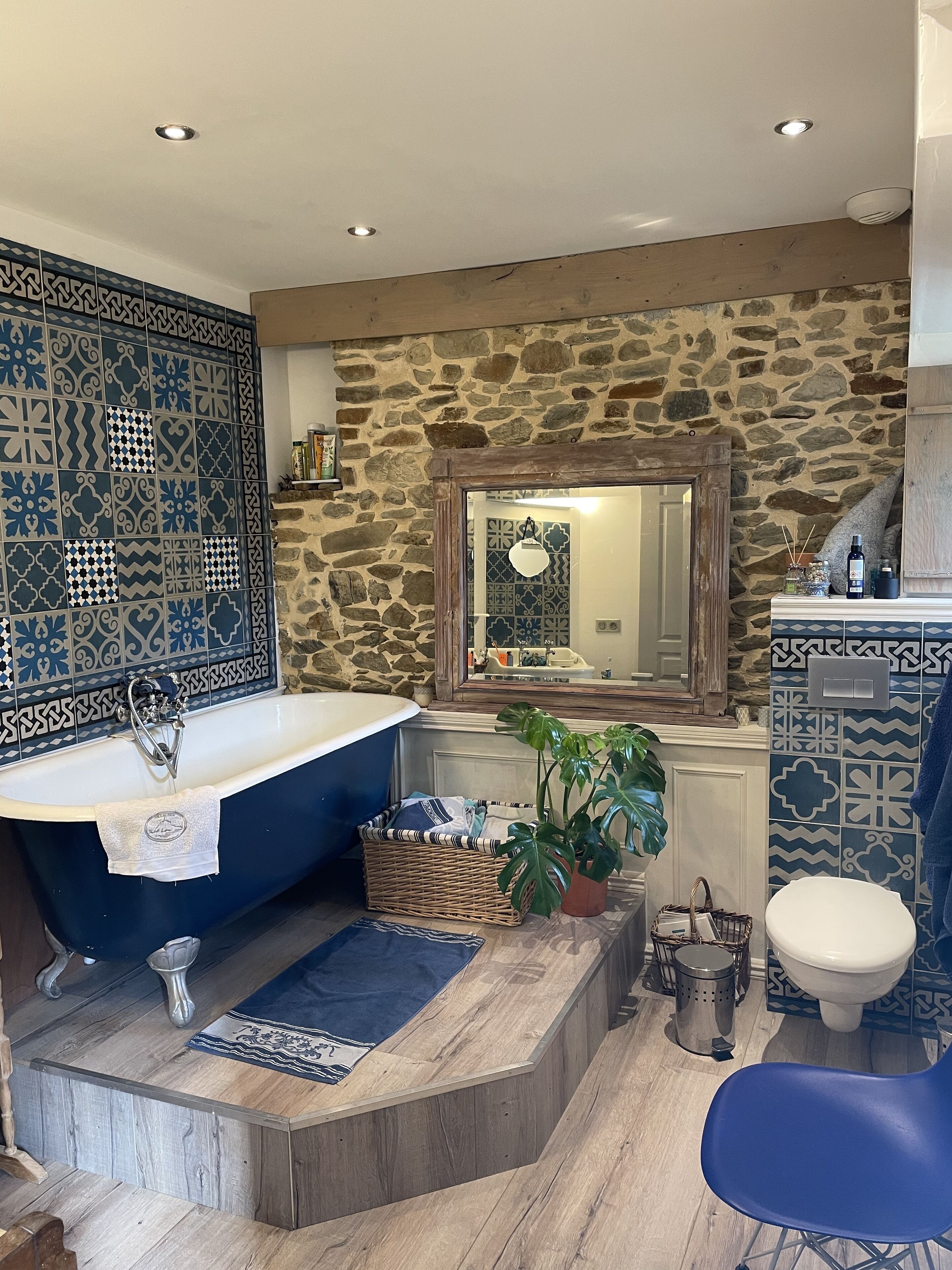 Une salle de bain avec une baignoire sur pieds, un mur en pierre, un miroir et une chaise bleue.