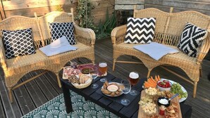 Two wicker chairs with patterned cushions, a wooden table with a variety of snacks and drinks, and a wooden deck with a green patterned rug.