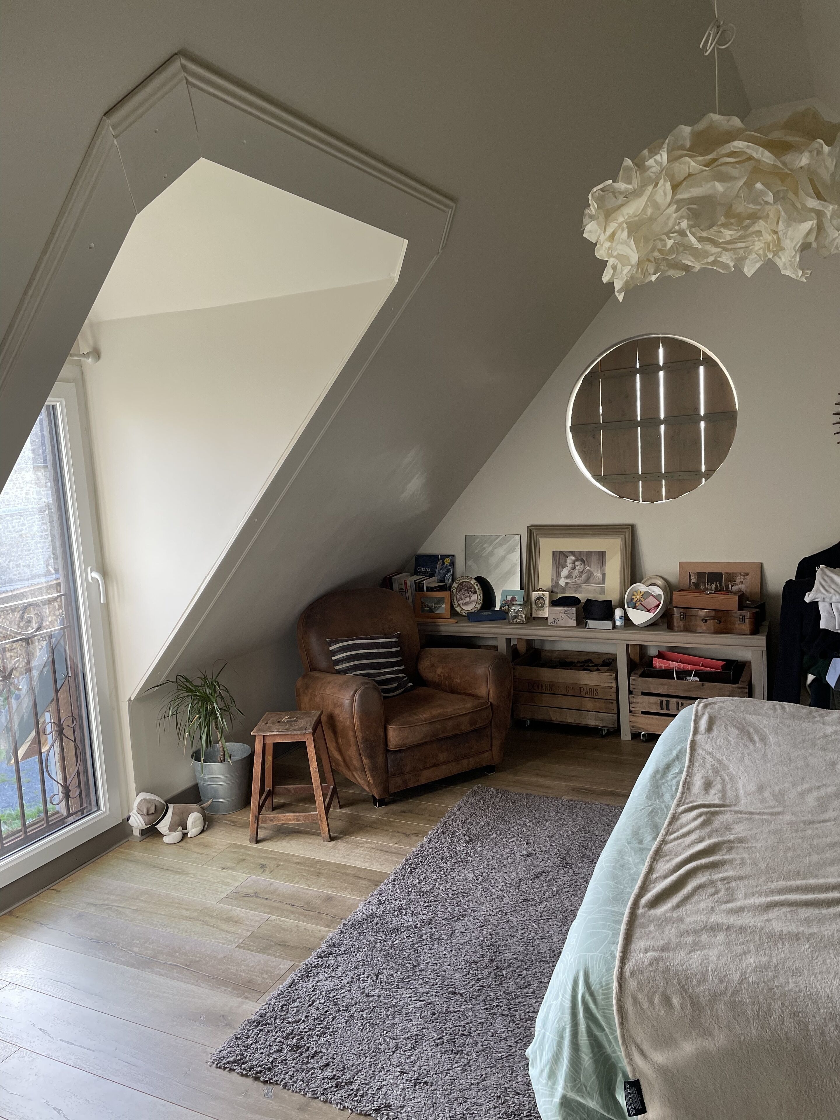 A cozy loft bedroom with a sloped ceiling, a large window, a brown leather chair, a small wooden table, a grey rug, and a round mirror on the wall.