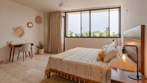 A bedroom with a large bed, a desk, and a chair. There is a window with a view of greenery outside. - Beautiful Residence with Rooftop & Small Pool  (Mérida)