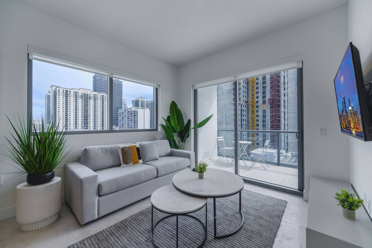 Ein modernes Wohnzimmer mit einem grauen Sofa, zwei runden Tischen und großen Fenstern mit Blick auf die Stadt.