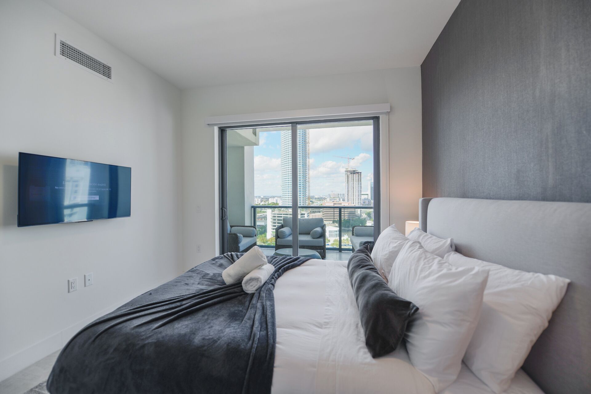A modern hotel room with a large bed, a flat-screen TV mounted on the wall, and a balcony with a city view.