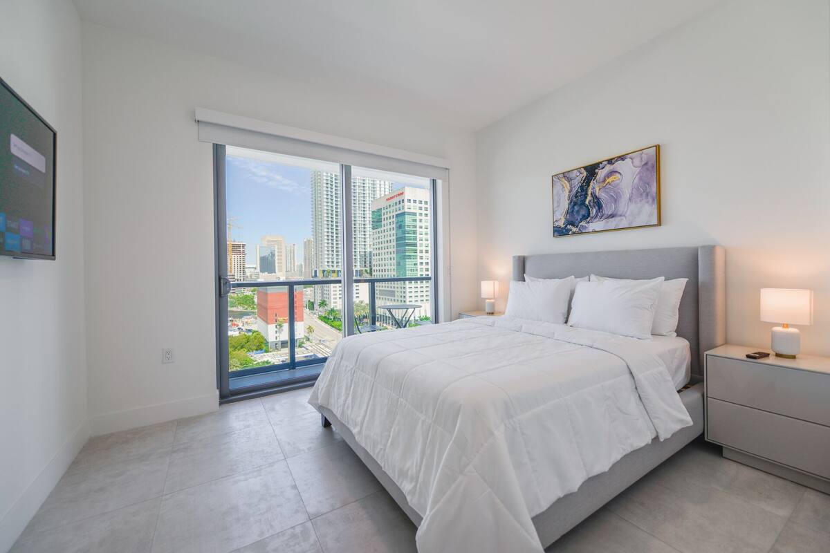 Ein modernes Hotelzimmer mit einem großen Bett, einem Nachttisch, einer Lampe, einem Bild an der Wand und Blick auf die Stadt durch ein großes Fenster.