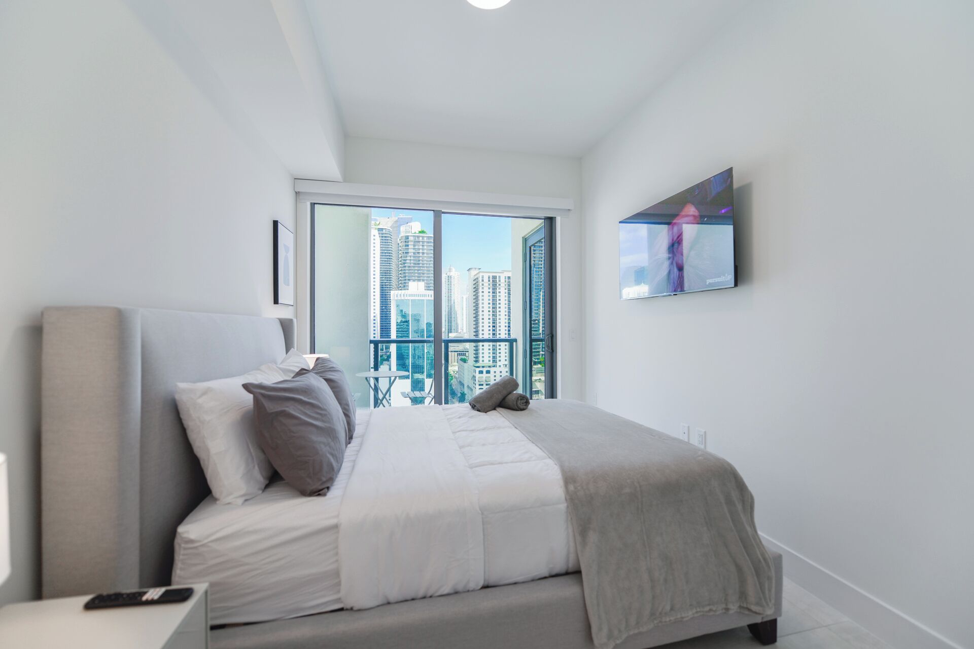 A modern bedroom with a large bed, a bedside table, a remote control, and a view of tall buildings through a large window.