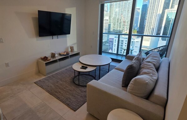 Ein modernes Wohnzimmer mit großem Fenster, Flachbildfernseher, rundem Couchtisch und beiger Couch.