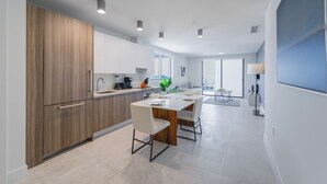 Une cuisine moderne avec des meubles en bois, un coin repas avec une table et des chaises blanches, et une vue sur la ville grâce à de grandes fenêtres.
