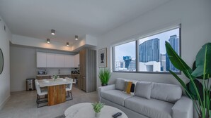 Un appartement moderne comprenant une cuisine, un coin repas et un salon. Il dispose d’une grande fenêtre offrant une vue sur la ville, d’un canapé gris et d’une table basse ronde.