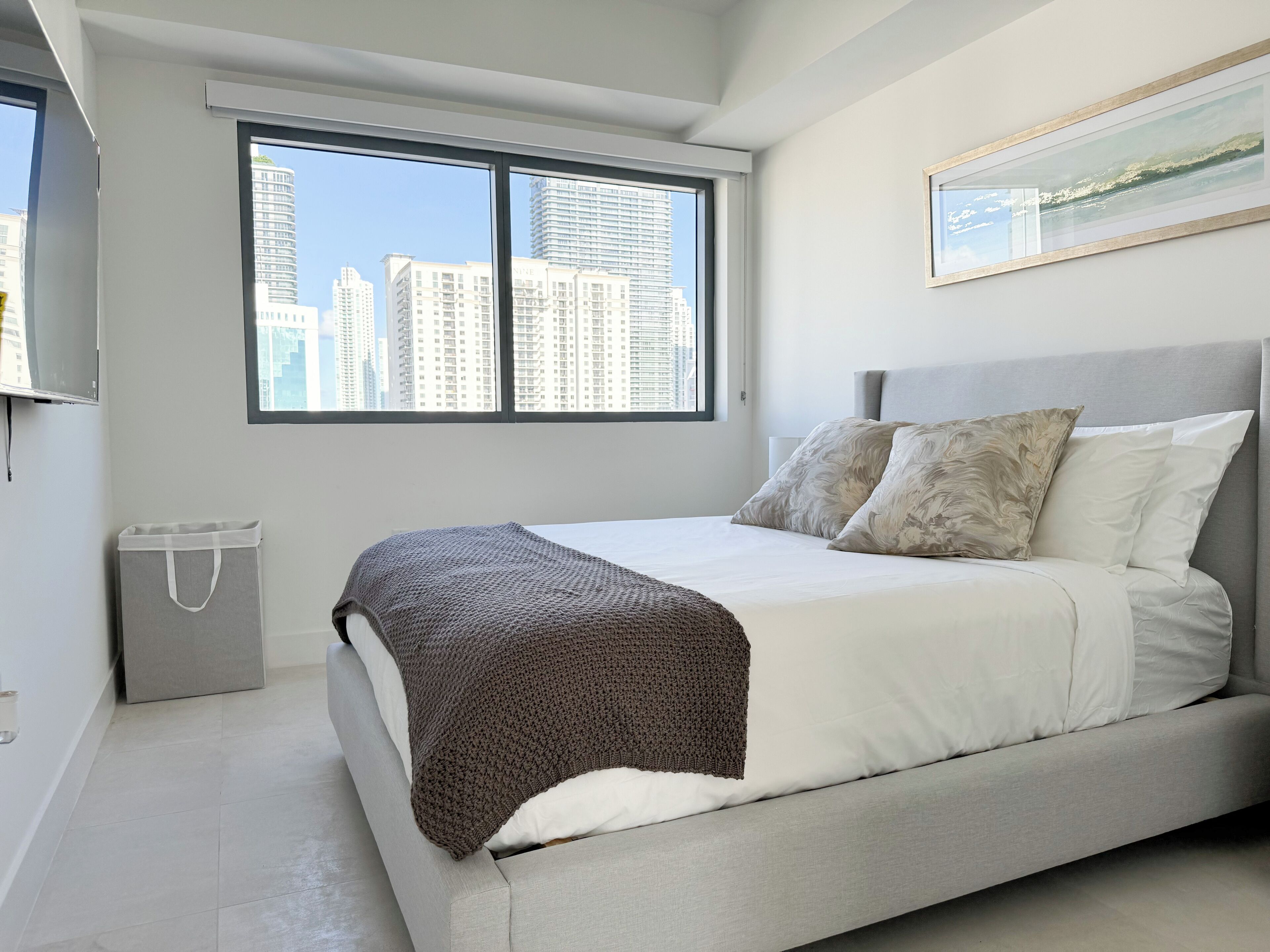 A modern bedroom with a large bed, a city view through the window, and a framed artwork on the wall.