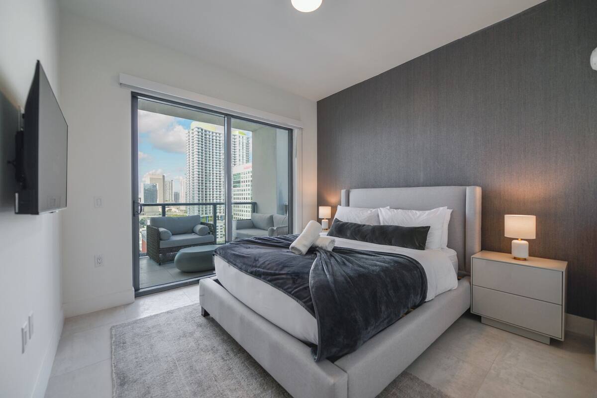 Ein modernes Hotelzimmer mit einem großen Bett, zwei Nachttischen mit Lampen, einem Sofa und einem Balkon mit Blick auf die Stadt.