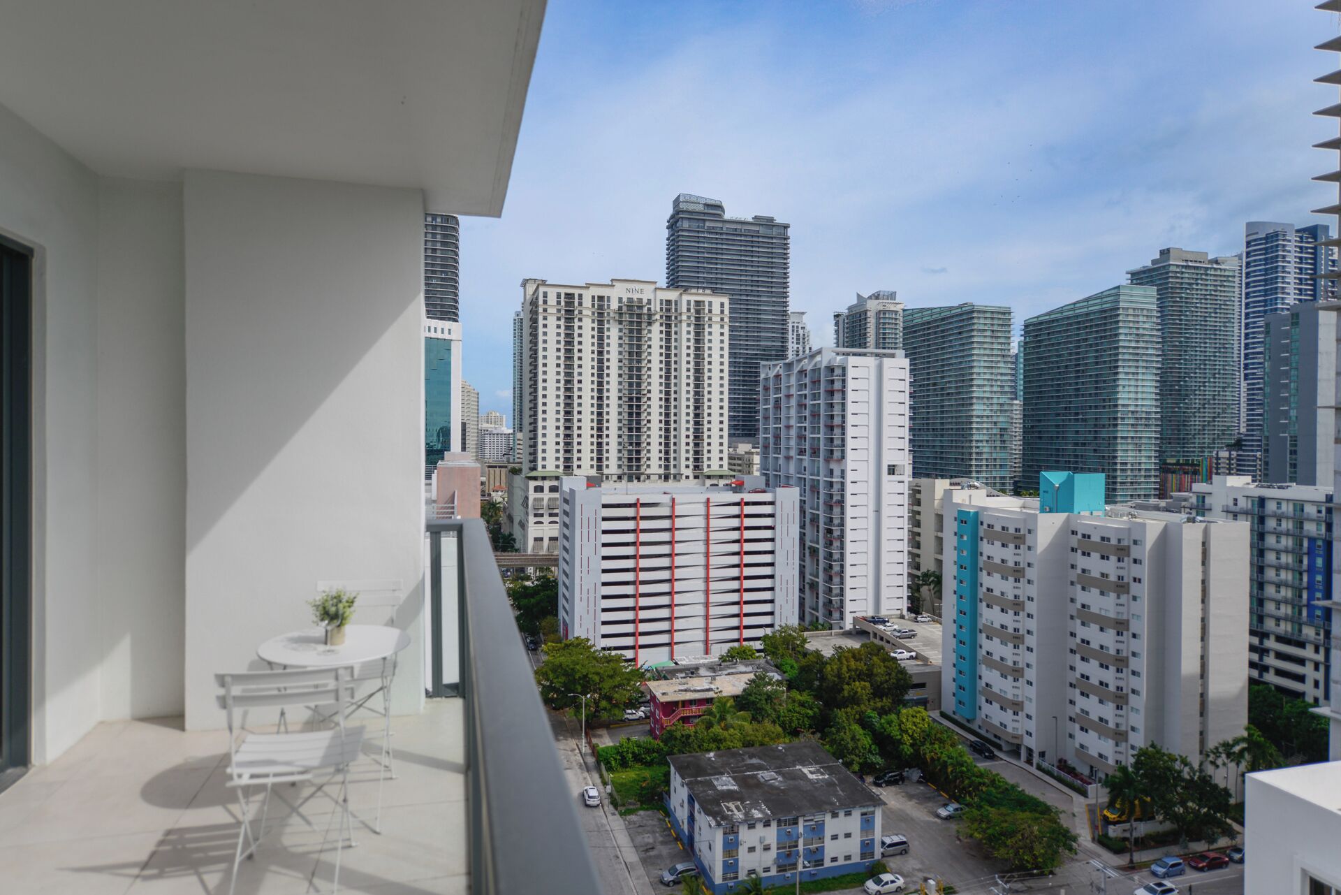 A balcony with a table and chairs, overlooking a cityscape with high-rise buildings and a parking area.