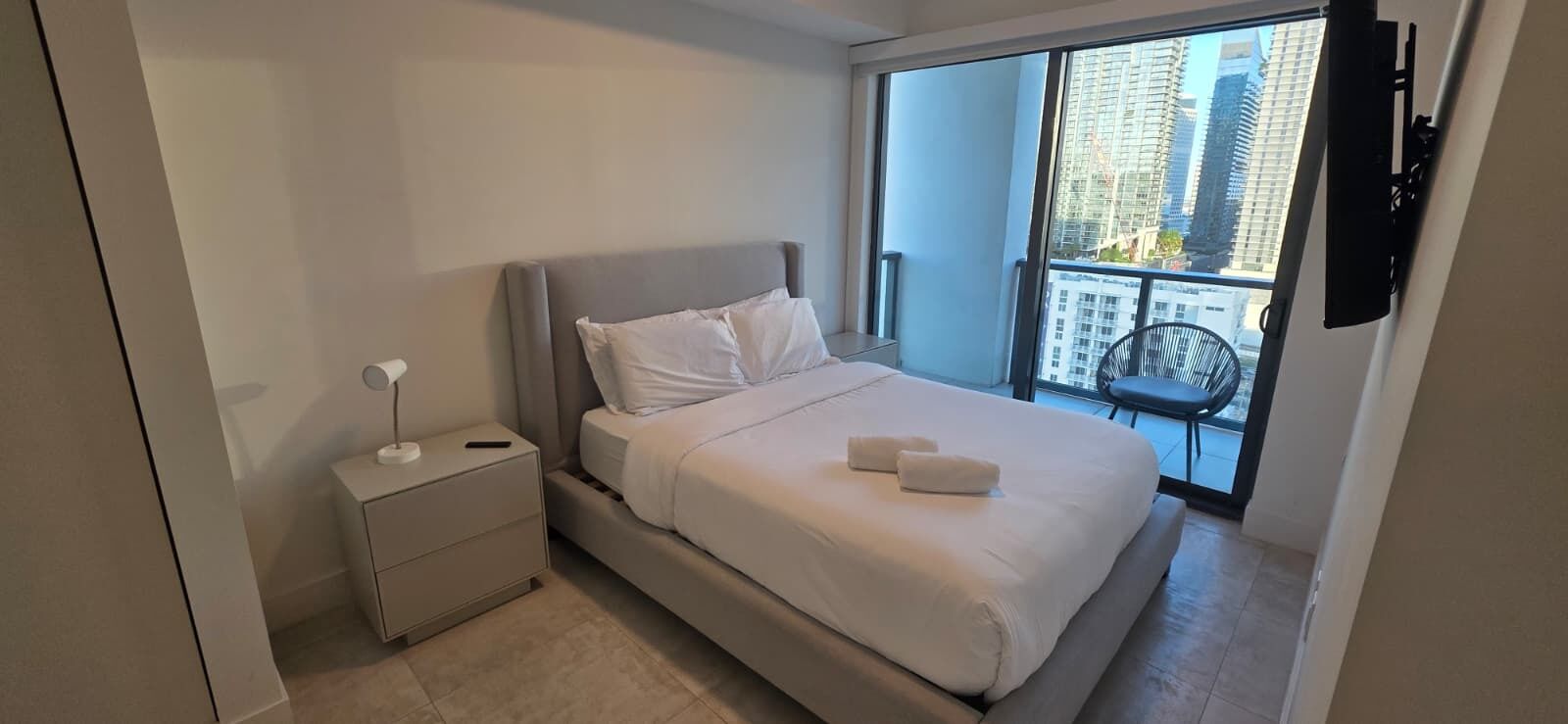 Un dormitorio moderno con cama, mesita de noche y vistas a la ciudad a través de un ventanal amplio.
