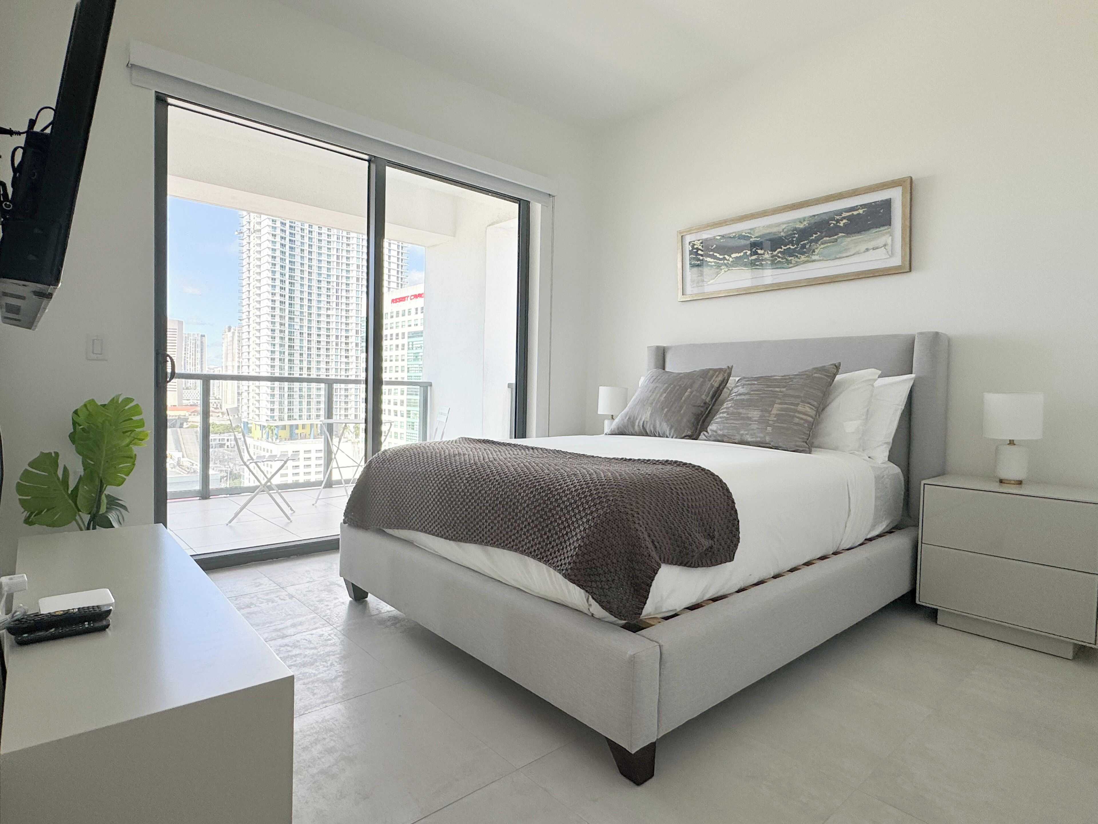 A modern bedroom with a large bed, a nightstand, a TV mounted on the wall, and a balcony with a city view.