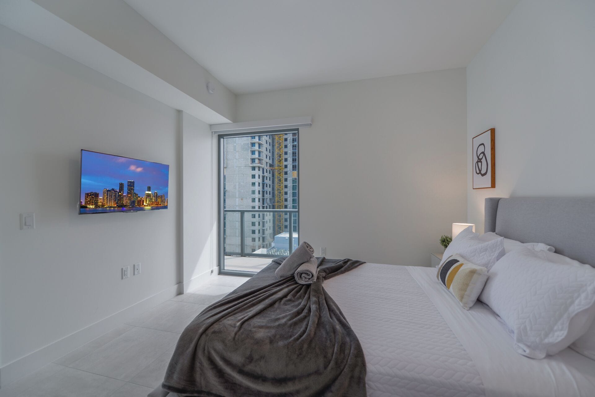 Una habitación de hotel moderna con una cama grande, un televisor de pantalla plana en la pared, un balcón con vista a la ciudad y una obra de arte abstracta enmarcada.