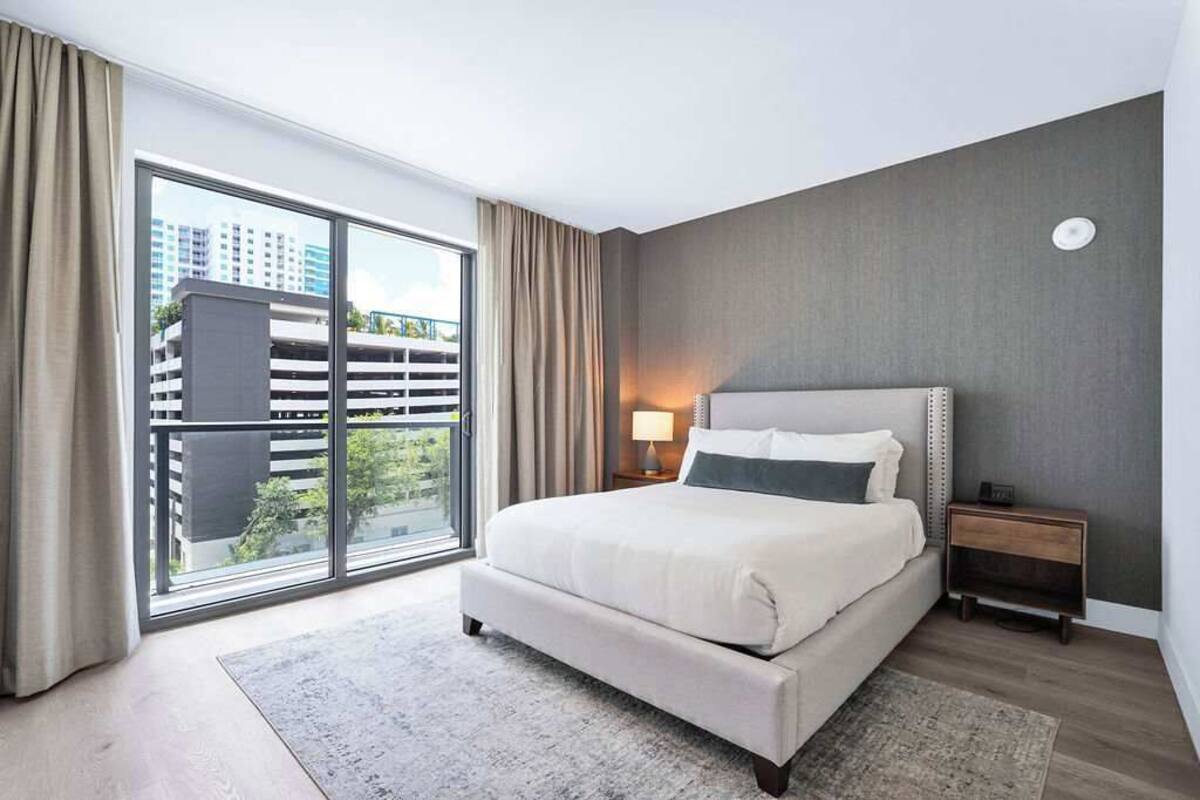 Ein modernes Hotelzimmer mit einem großen Bett, einem Nachttisch, einer Lampe und Blick auf eine Parkgarage durch große Fenster.